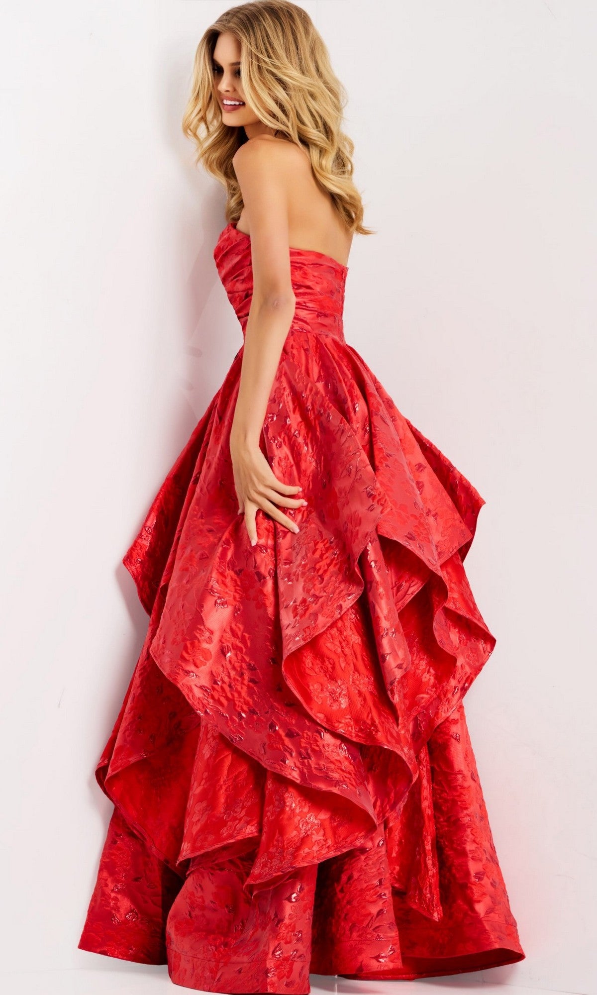A woman wearing the Long Prom Dress: Jovani 47394, a strapless bright red gown with a ruffled layered skirt, stands against a plain white background.